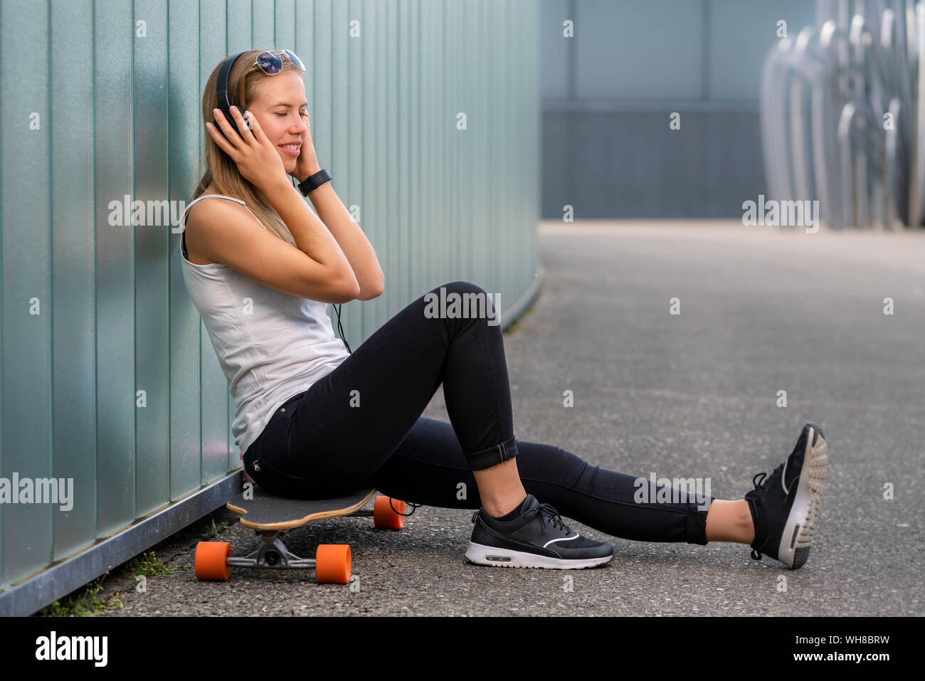 Lächelnde junge Frau sitzt auf LONGBOARD Musik hören mit Kopfhörern Stockfoto