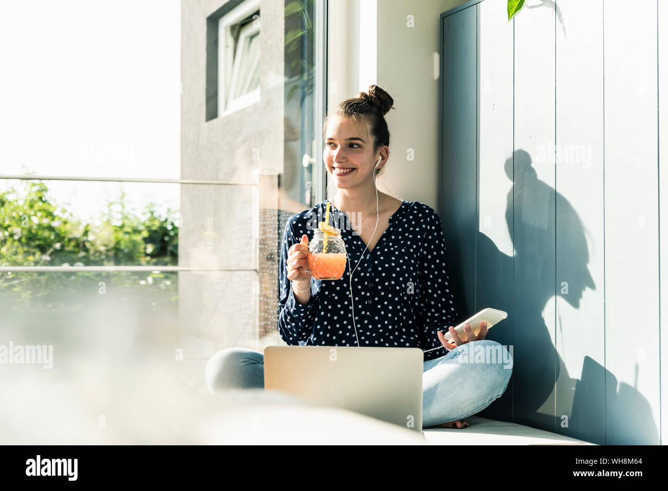 Lächelnde junge Frau mit Laptop, Handy und gesundes Getränk zu Hause Stockfoto