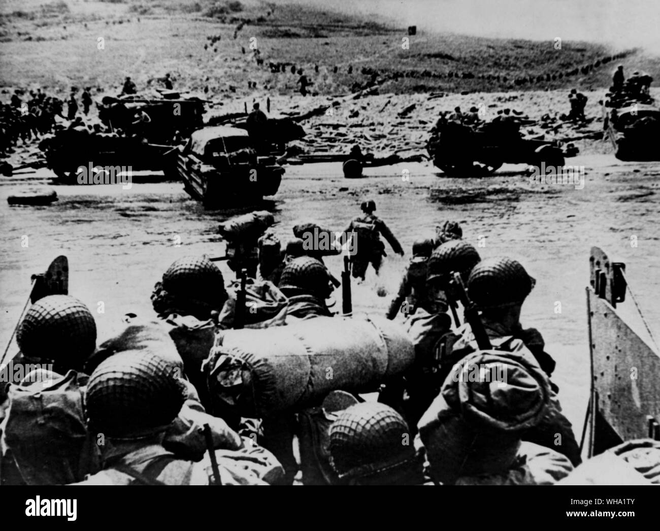 WW2: D-Tag. Amerikanische stoßtruppen von einem Landing Craft durch das flache Wasser auf einen Strand - Kopf im nördlichen Frankreich während der Landung der Alliierten. Hälfte - Titel und ein Strände DUKW am Strand - Kopf. Der Rauch im Hintergrund ist von naval Geschützfeuer Unterstützung der Angriff. 6. Juni 1944. Stockfoto