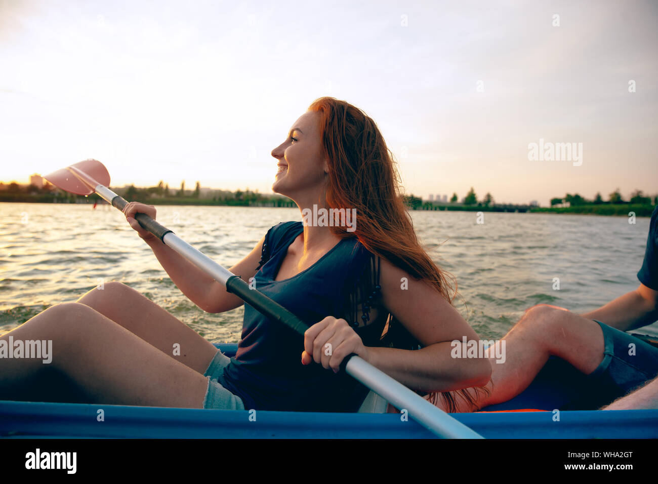 Selbstbewussten jungen kaukasischen Frau Kajakfahren auf dem Fluss mit Sonnenuntergang im Hintergrund. Spaß in der Freizeit. Gerne Haar weiblichen Modell auf dem Kajak. Sport, Beziehungen Konzept. Stockfoto