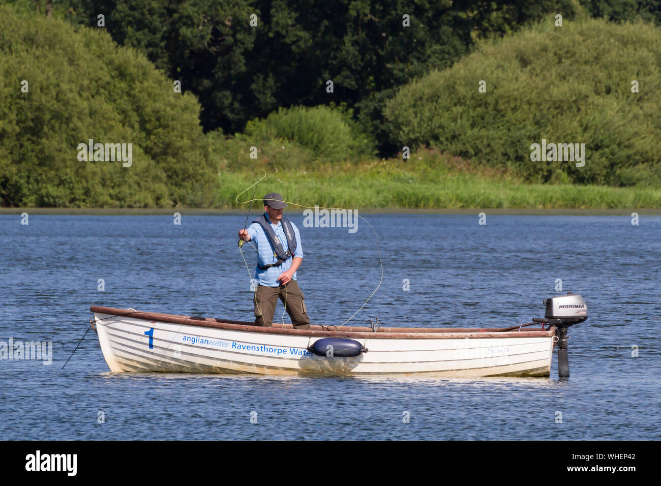 Der "Ravensthorpe Reservoir" in Northamptonshire, Großbritannien: Ein Angler mit flacher Kappe steht in einem kleinen Boot mit Außenbordmotor und wirft seine Linie. Stockfoto