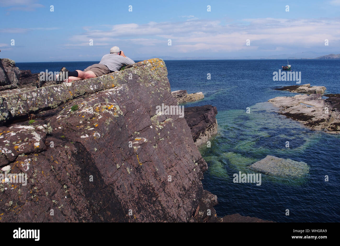 Ein älterer Mann mit dem Gesicht nach unten auf den Felsen, tiefe blaue Meer mit dem Fernglas suchen und trug eine Kappe und Kurzschlüsse in der Nähe von Culkein, Sutherland, Schottland Stockfoto