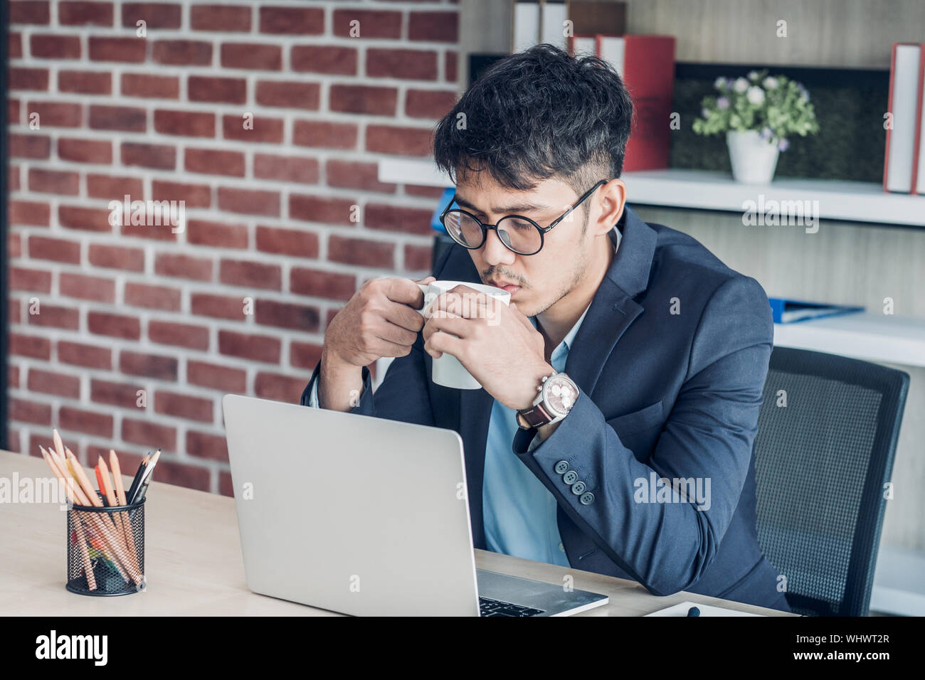 Junge asiatische Geschäftsmann tragen Anzug trinken Kaffee und siehe Bildschirm während der Arbeit mit dem Laptop auf dem Schreibtisch Tisch in modernen Büro. Anschließen des Network online conce Stockfoto