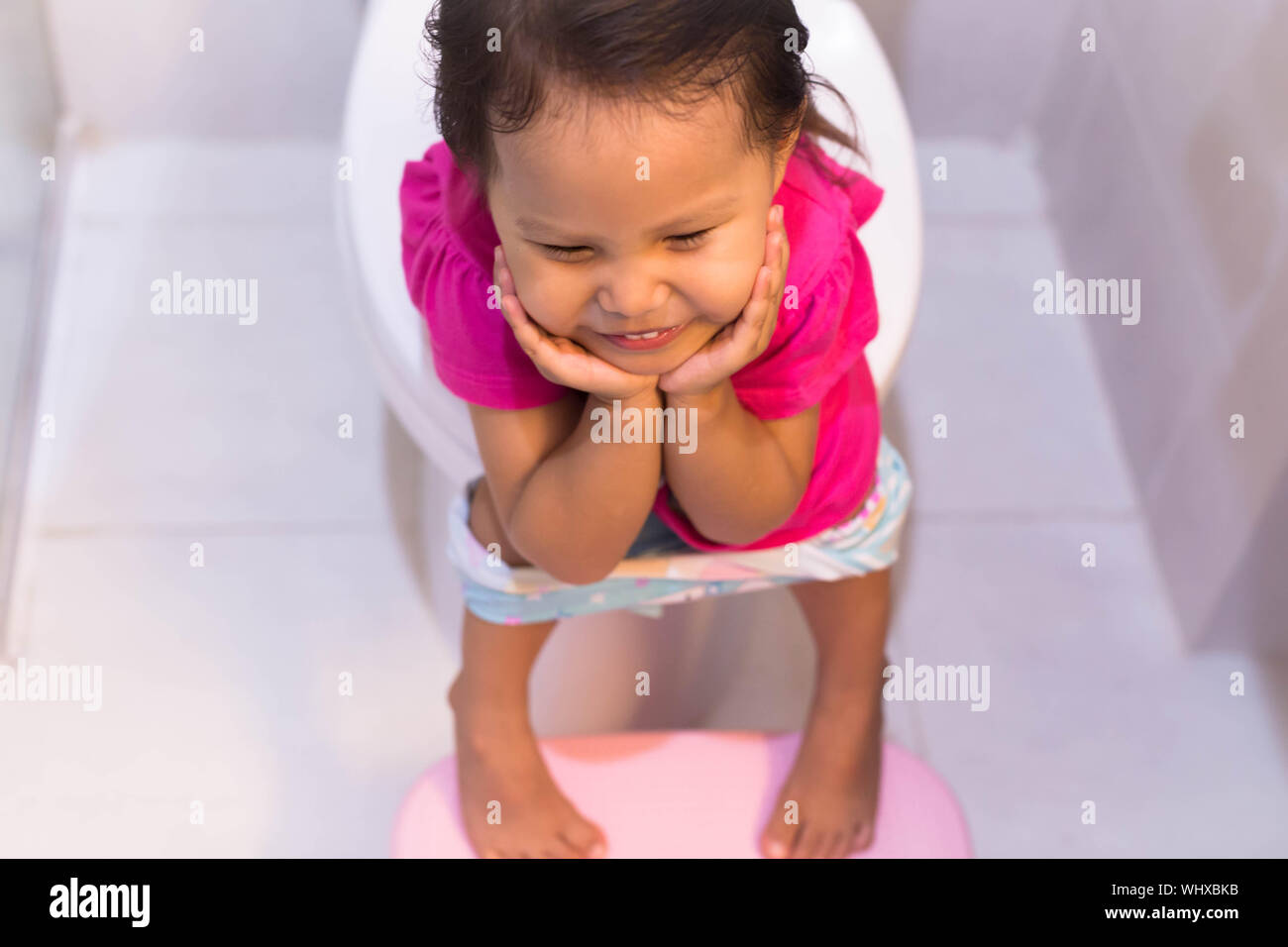 Ein wenig weibliches Kind auf der großen Toilette sitzt in einem weißen Badezimmer, lächelnd und versuchen, Pinkeln zu gehen. Stockfoto