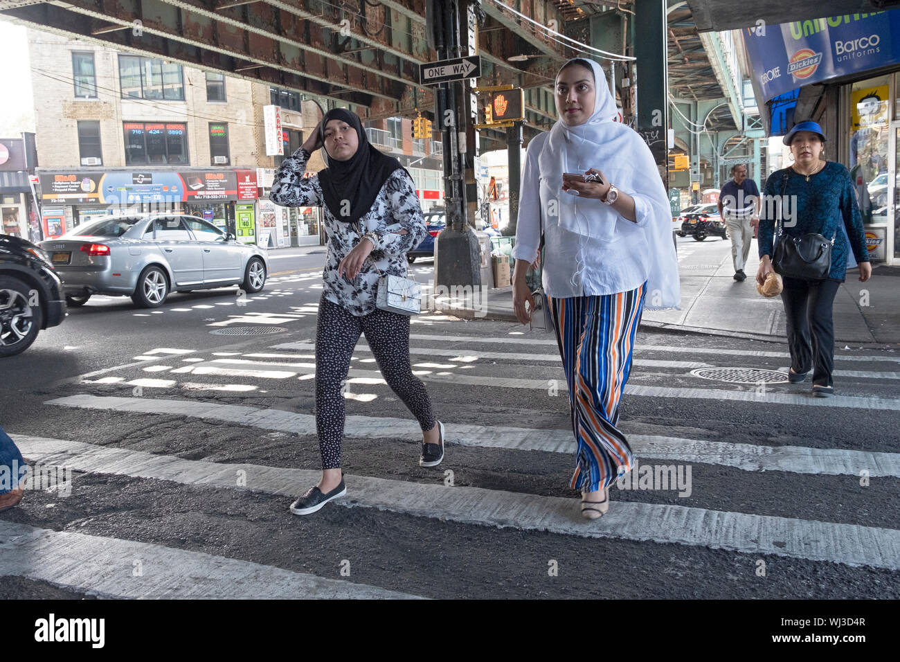 Zwei elegant Kleider Frauen in hijabs Überqueren einer Straße auf Roosevelt Ave. Unter der erhöhten U-Bahn. Jackson Heights, Queens, New York City. Stockfoto