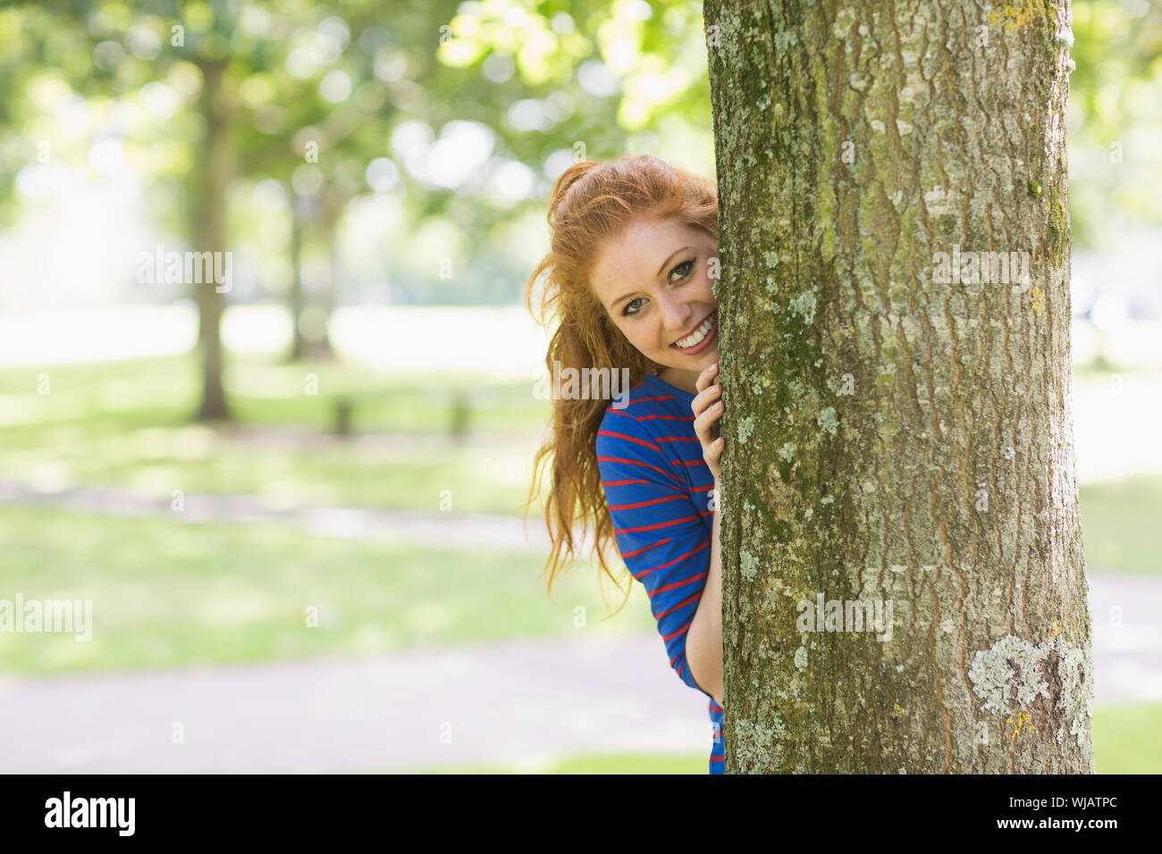 Lächelnde hübsche Rothaarige versteckt sich hinter einem Baum Stockfoto