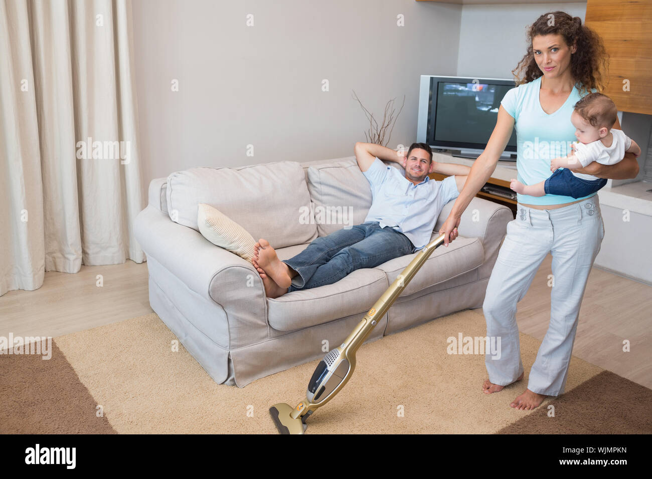 In voller Länge Porträt von Frau Reinigung Haus, während Baby mit man entspannt auf einem Sofa Stockfoto