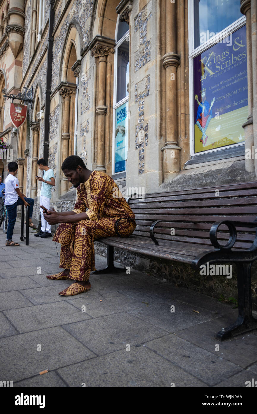 Winchester, Hampshire, UK ein schwarzer Mann mit traditionellen afrikanischen Kleid auf einer Bank sitzen, auf sein Handy oder Mobiltelefon suchen Stockfoto