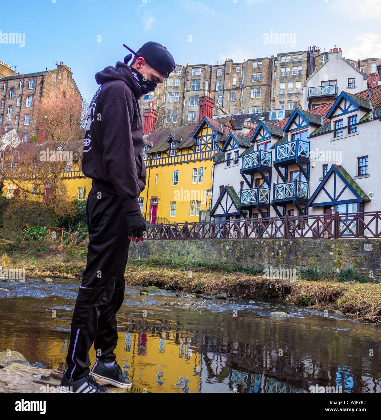 Eine Person, die einen Hoodie und eine schwarze Mütze trägt Ein schmutziger Teich in der Nähe von Gebäuden und Häusern Stockfoto