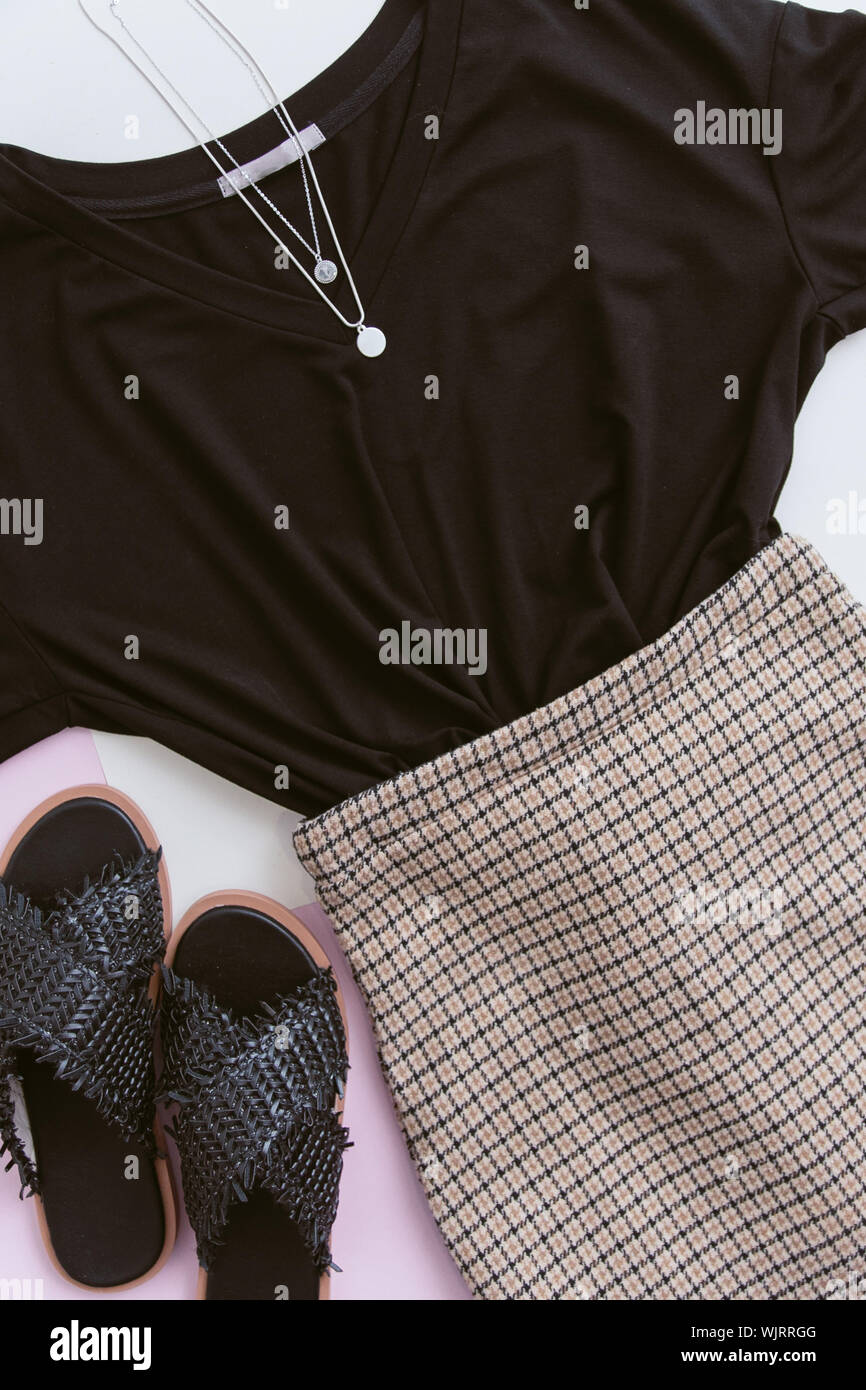 Lässig auf einem Pastell rosa Hintergrund. schwarzes T-Shirt, karierten rock und Silber Anhänger. schwarz Stroh Sandalen. Sommer Konzept der Frauen s trend Kleider und Schuhe Stockfoto