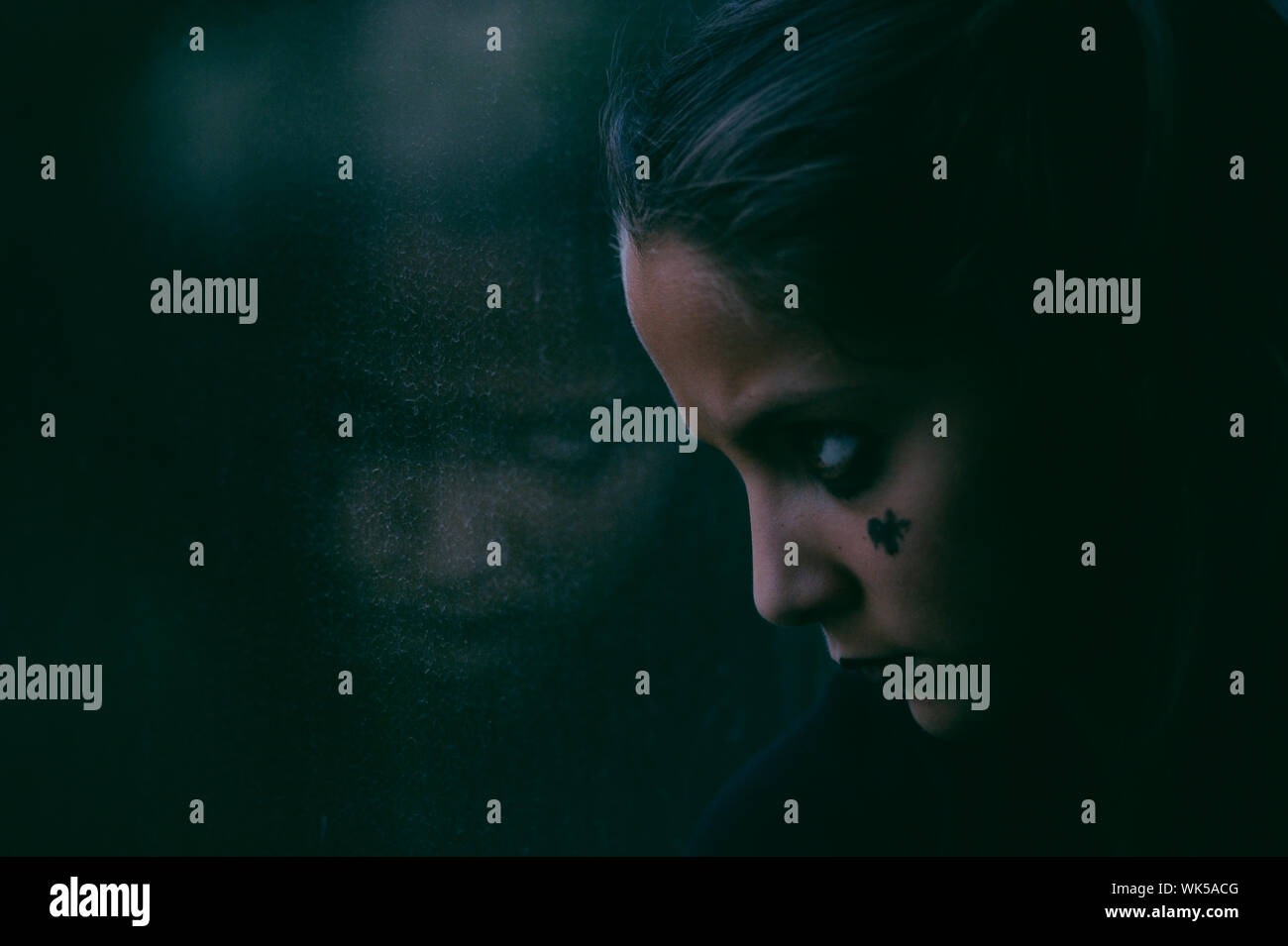 Junge Mädchen mit dunklem Makeup für Halloween Party auf ihr Spiegelbild im Fenster beleuchtet Film grünlichen Licht kaufen Stockfoto
