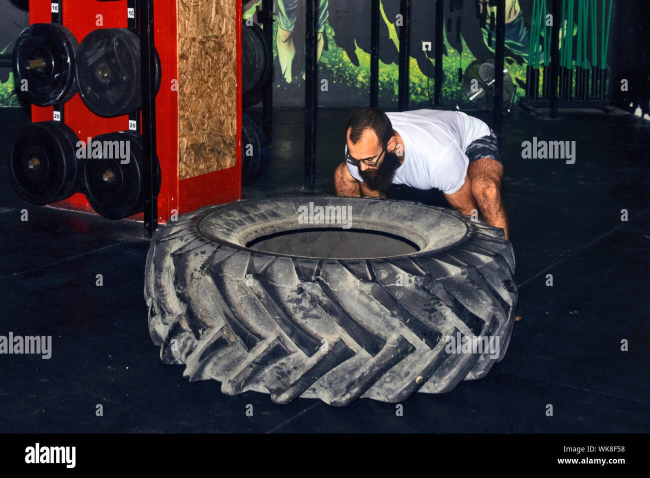 Ein Mann ist in der Fitness, dreht ein großes Rad von einem Traktor auf dem Boden einer Sporthalle. Stockfoto