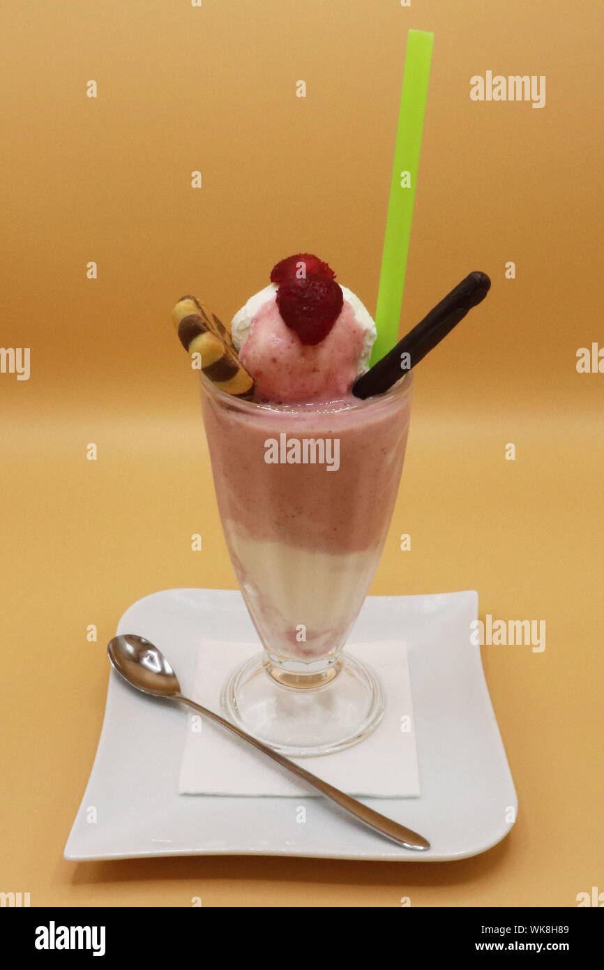 Hohes Glas Erdbeeren- und Creme-Milchshake mit Himbeer- und Waffelstäbchen, serviert mit grünem Stroh und einem Löffel auf einem weißen Teller Stockfoto