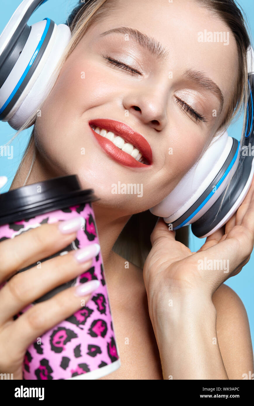 Junge ziemlich glücklich Frau trinkt Kaffee und das Hören von Musik über Kopfhörer. Frau mit geschlossenen Augen auf blauem Hintergrund. Lächelnde Mädchen mit Kaffee pa Stockfoto