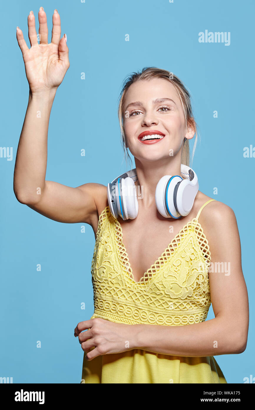 Junge hübsche Frau Hören von Musik über Kopfhörer. Frau im gelben Kleid winkende Hand auf blauem Hintergrund. Lächelnde Mädchen mit großen Kopfhörer auf Stockfoto