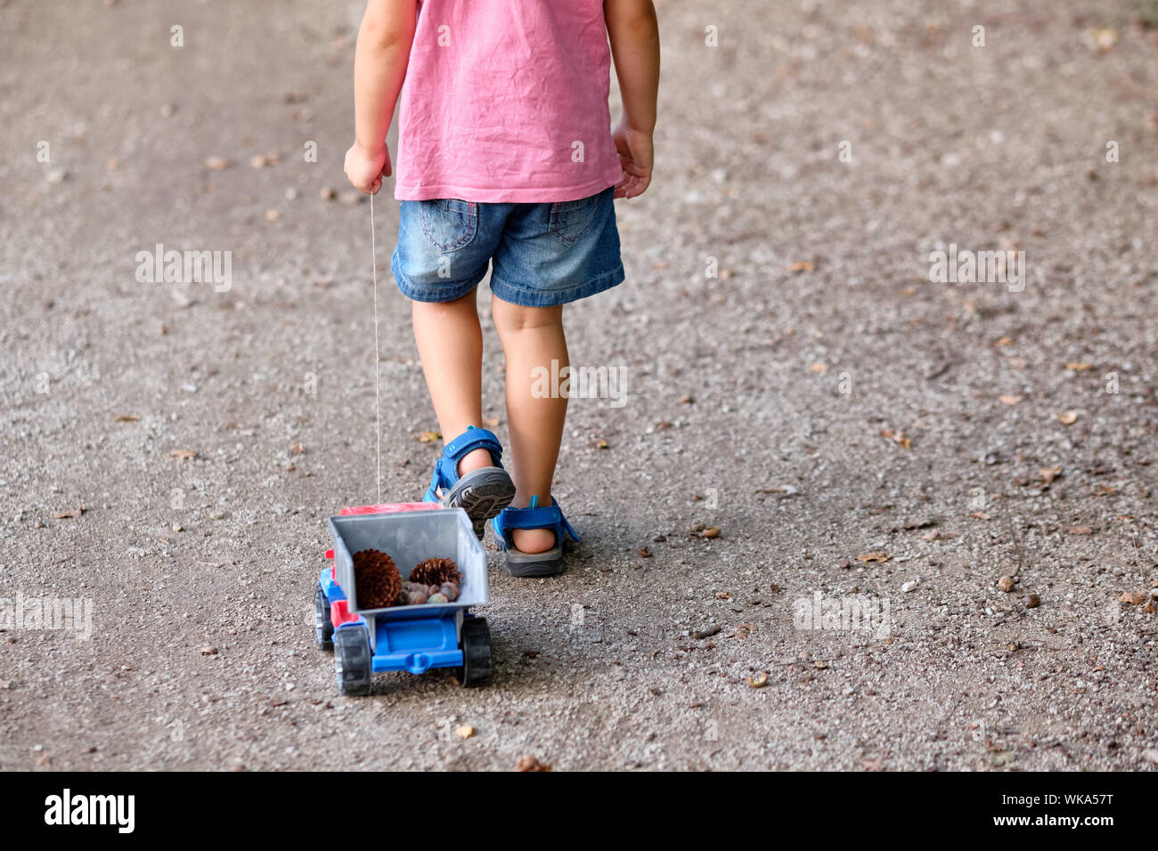 Rückansicht des unteren Abschnitt von einem 3-4 jährigen Kind im Sommer Kleidung auf einer Kiesgrube Fußweg Ziehen einer Spielzeug-LKW mit Kegel in Es Stockfoto