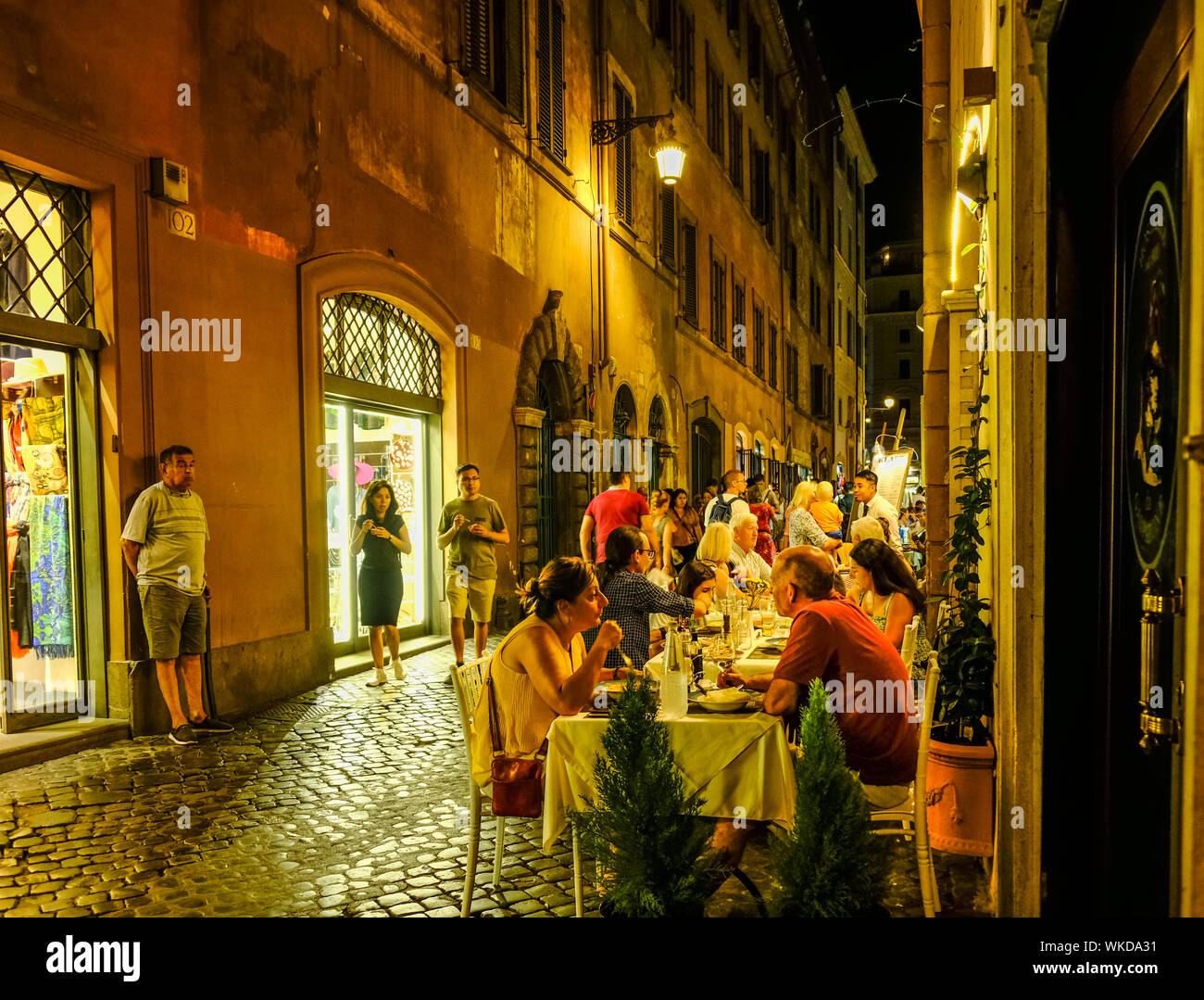 Nacht Blick auf die schmale Straße in Rom, Italien; speichert auf der linken und Outdoor Restaurant mit Menschen essen auf der rechten Seite Stockfoto