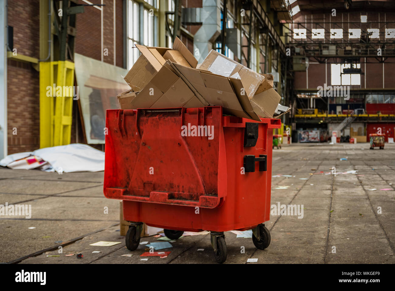 Überquellenden Mülleimer in der Mitte einer großen Leere Industriehalle sehr rot mit grün gelb und schwarz Lack auf Rädern Stockfoto