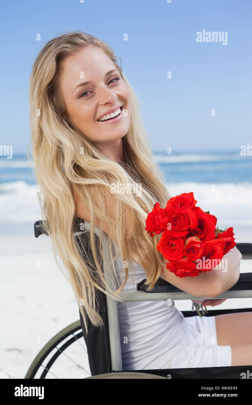 Rollstuhl blond lächelnd in die Kamera am Strand halten Rosen an einem sonnigen Tag gebunden Stockfoto
