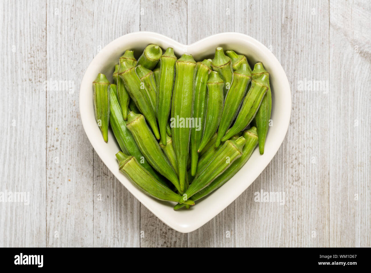 Frische organische Grün okra in Herzform Schüssel auf ein weißes Holzbrett isoliert. Stockfoto