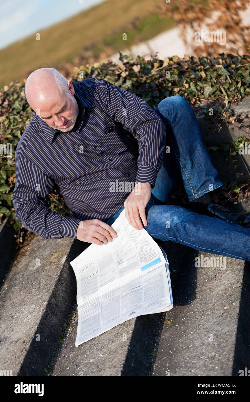 Glatze Mann mittleren Alters in Jeans sitzen im Freien in der Sonne auf einer Treppe eine Zeitung lesen Stockfoto