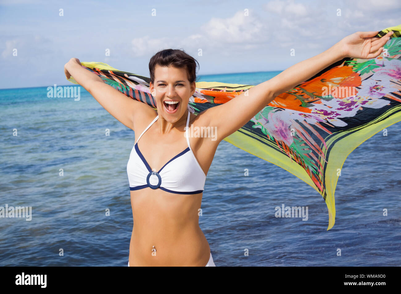 Lachend temperamentvolle Frau im Bikini am Meer mit farbenfroh gemusterten Schal in ihrem ausgestreckten Händen in der Brise gegen ein Flattern Stockfoto