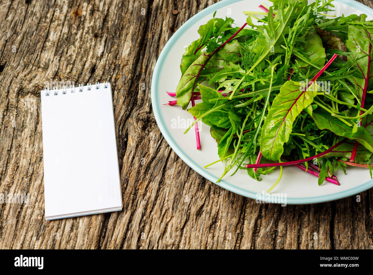 Frischer Salatteller mit gemischtem Grün (Rucola, Mesclun, Mache) auf dunklem Holzhintergrund mit Notizblock aus nächster Nähe. Gesunde Ernährung. Grüne Mahlzeit. Flach liegend Stockfoto