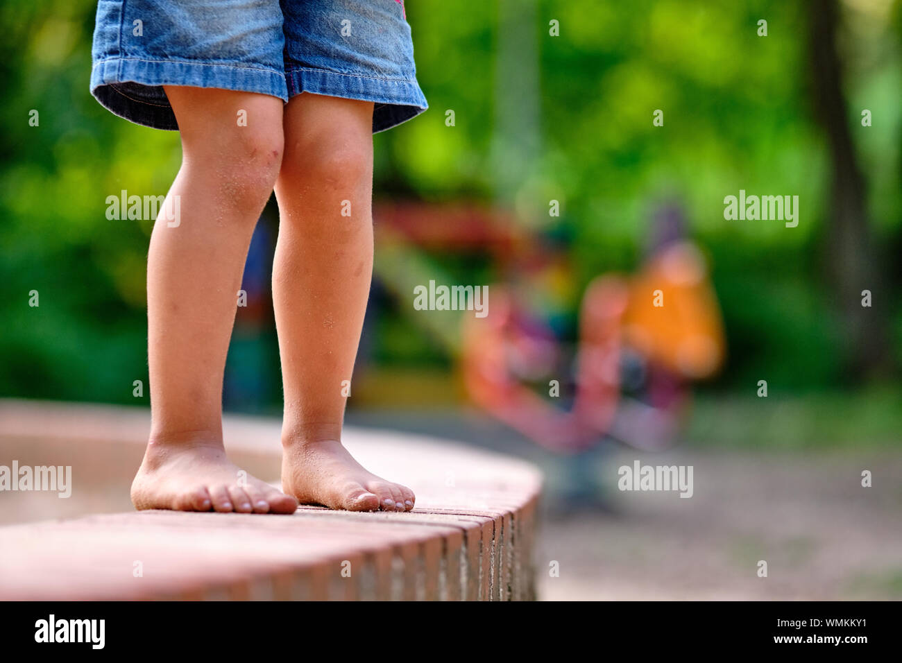 Niedrige Abschnitt einer kaukasischen Kind Mädchen in Jeans Shorts, die Barfuß steht auf einer kleinen Steinmauer mit sandigen Füßen im Sommer Stockfoto