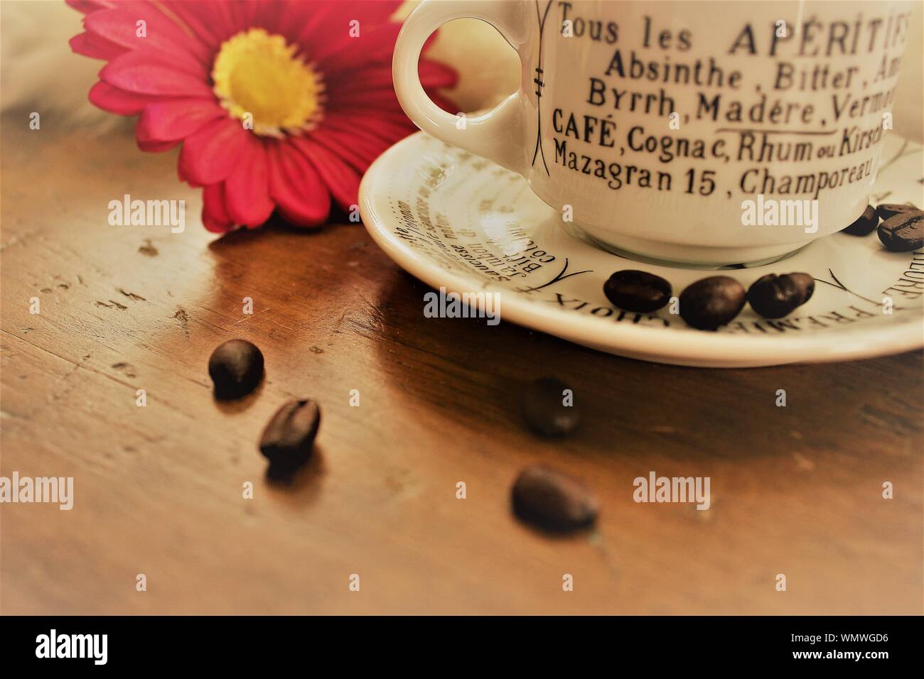 Eine kleine weiße Tasse mit Aperitifs in französischer Sprache aufgeführt ist, auf einen hölzernen Tisch, mit Kaffeebohnen und eine rosa Blume neben ihm. Stockfoto