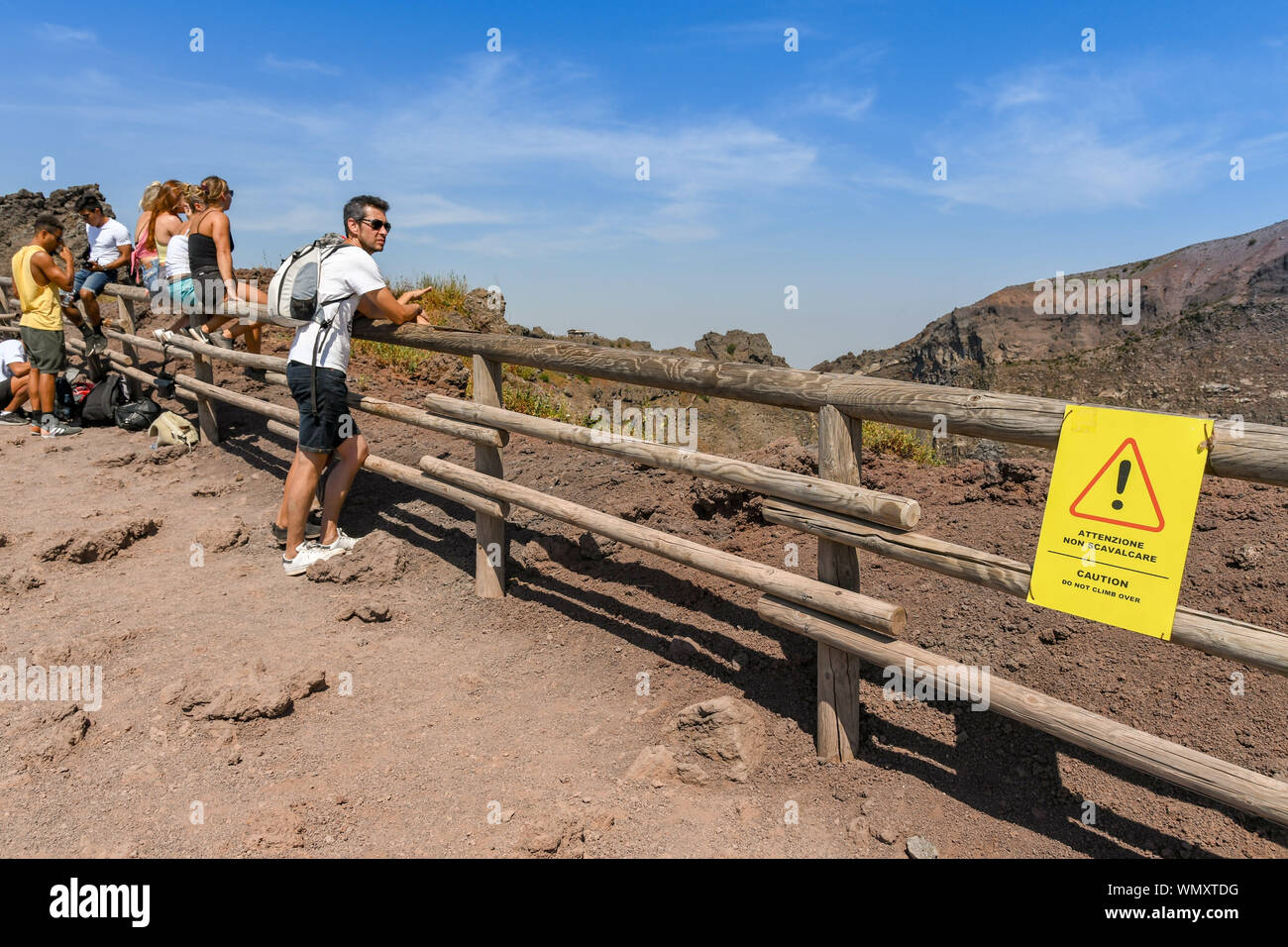 Neapel, Italien - AUGUST 2019: Menschen sitzen und lehnte sich auf den Zaun rund um den Krater des Vesuvs. Auf der rechten Seite ist ein Warnzeichen Stockfoto