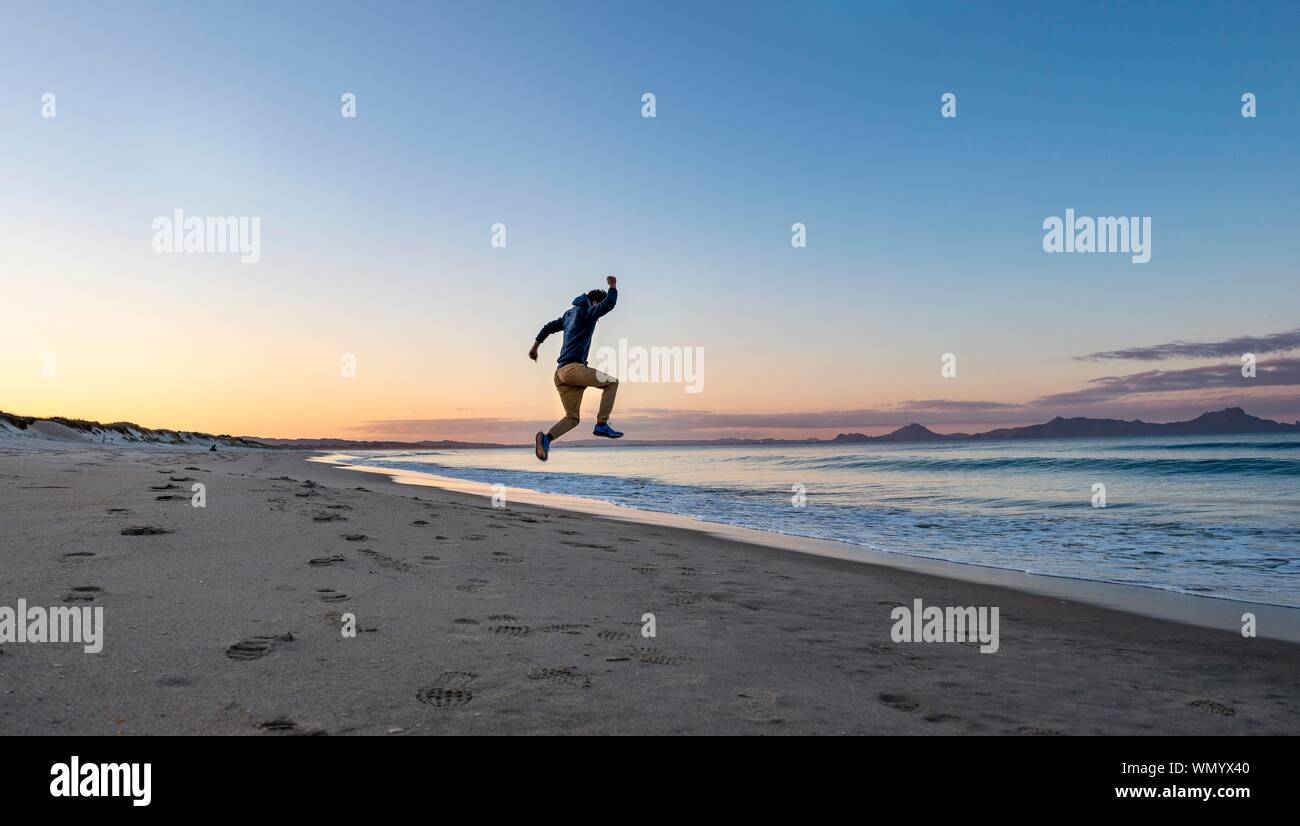 Junger Mann springt in die Luft, Strand Waipu Strand bei Sonnenuntergang, waipu Cove, Northland, Neuseeland Stockfoto