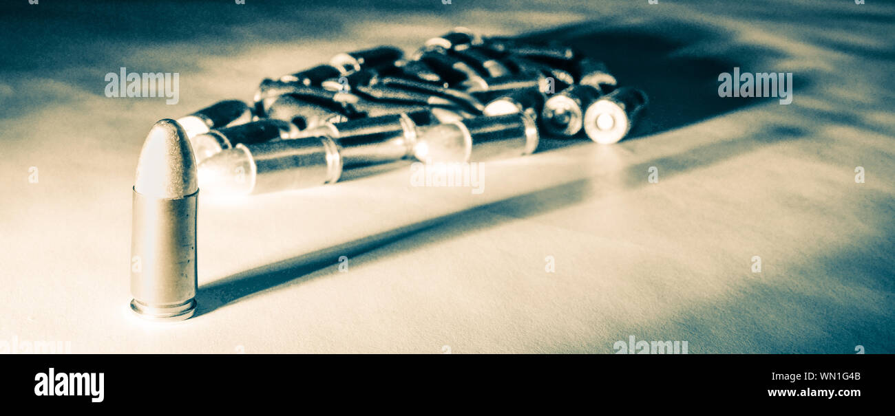 Eine einzelne 9mm bullet steht allein mit starken seitlichen Licht erzeugen lange Schatten mit einem Stapel von Kugeln im Hintergrund, im grünlichen Ton Schwarz und Weiß Stockfoto