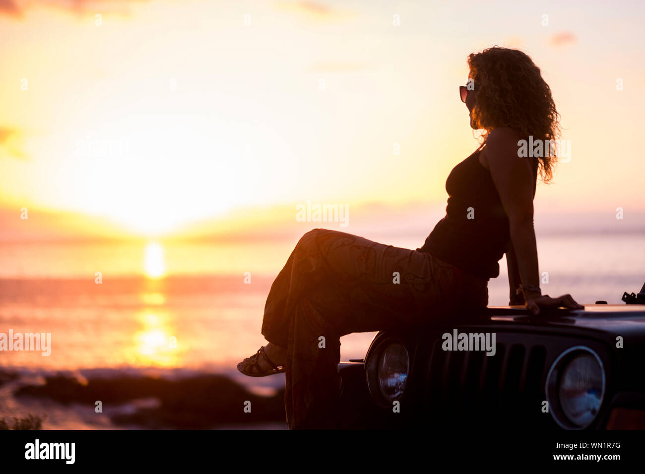 Frau sitzt auf dem Auto mit Strand bei Sonnenuntergang Stockfoto