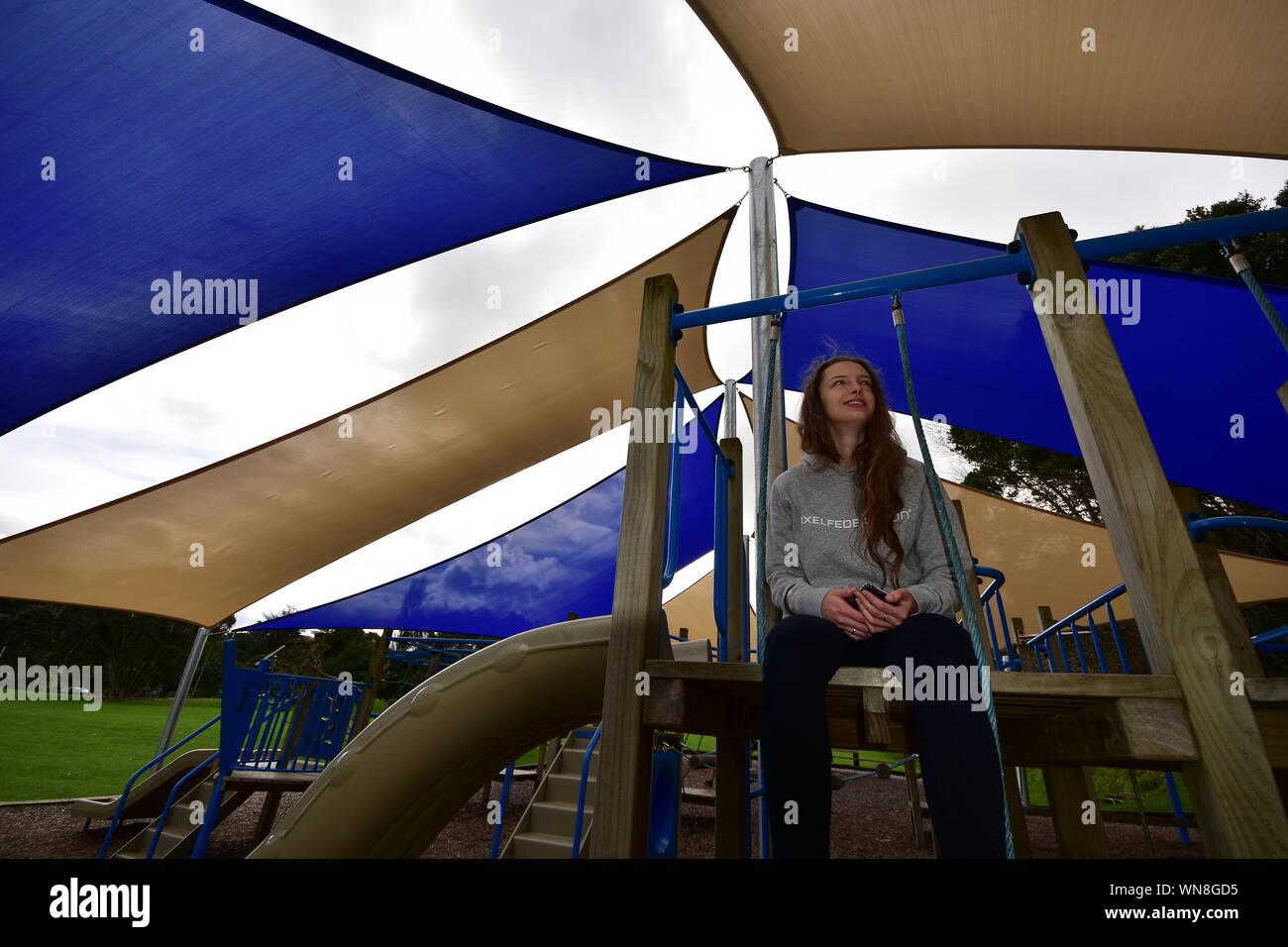 Mädchen mit langem Haar sitzt in der Nähe der Rutschbahn unter Schatten Segel an bewölkten Tag. Stockfoto