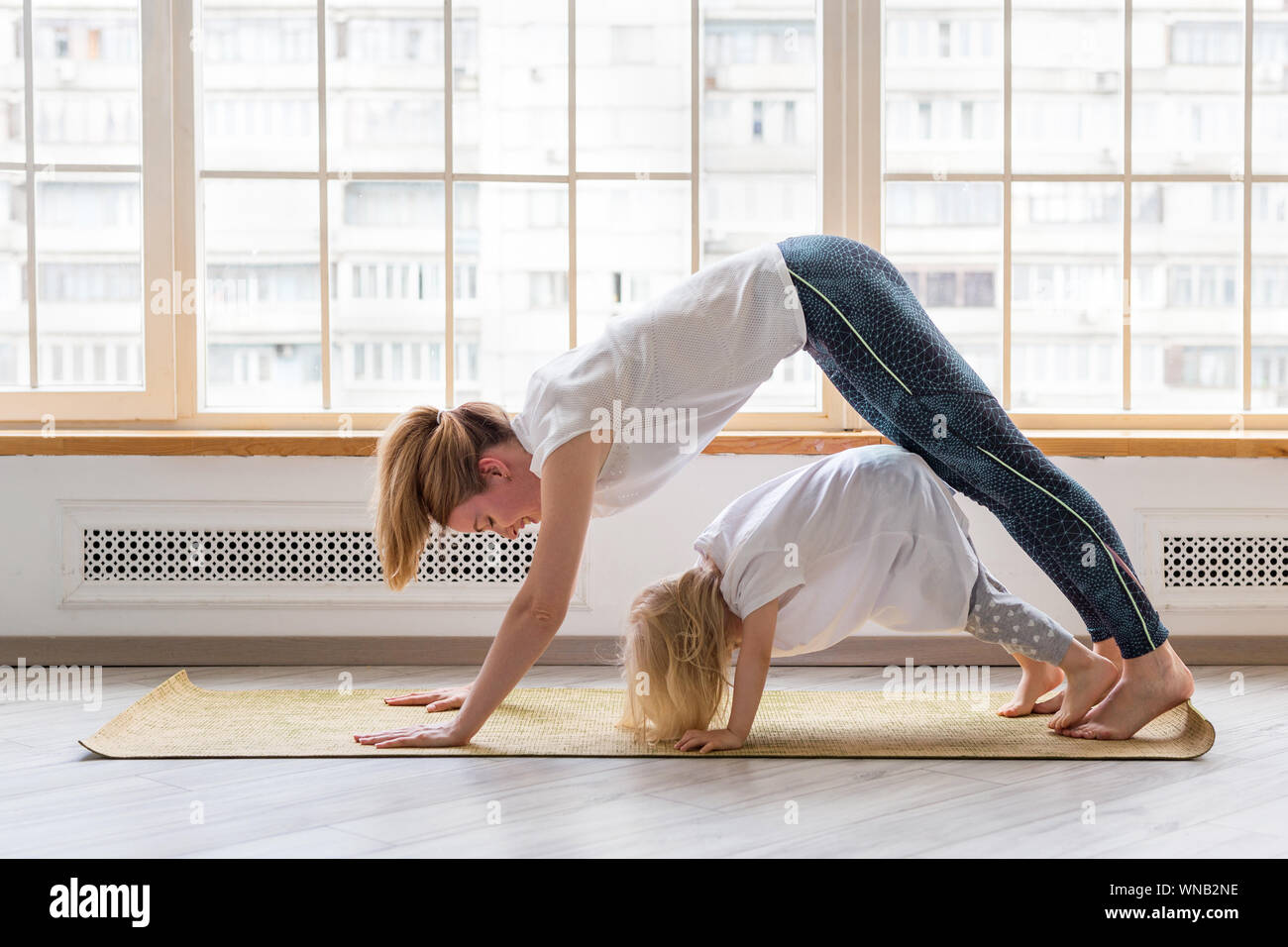 Junge Mutter macht Yoga mit 3-jährigen Mädchen vor dem Fenster. Nach unten schauender Hund Asana Stockfoto