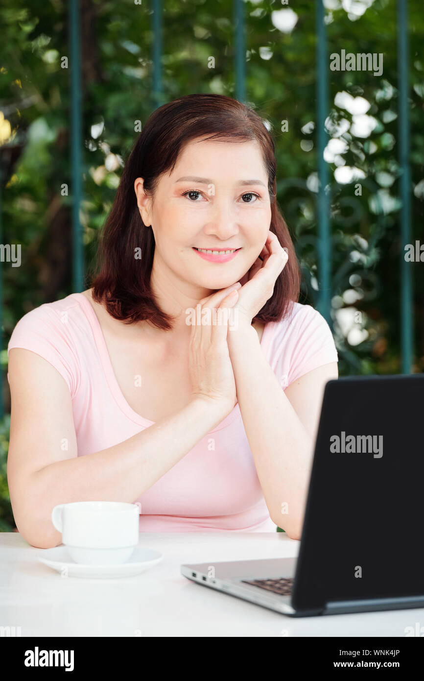 Portrait von positiven reife weibliche Freiberufler sitzen im Cafe Tabelle mit Tasse Kaffee und Arbeiten am Laptop Stockfoto