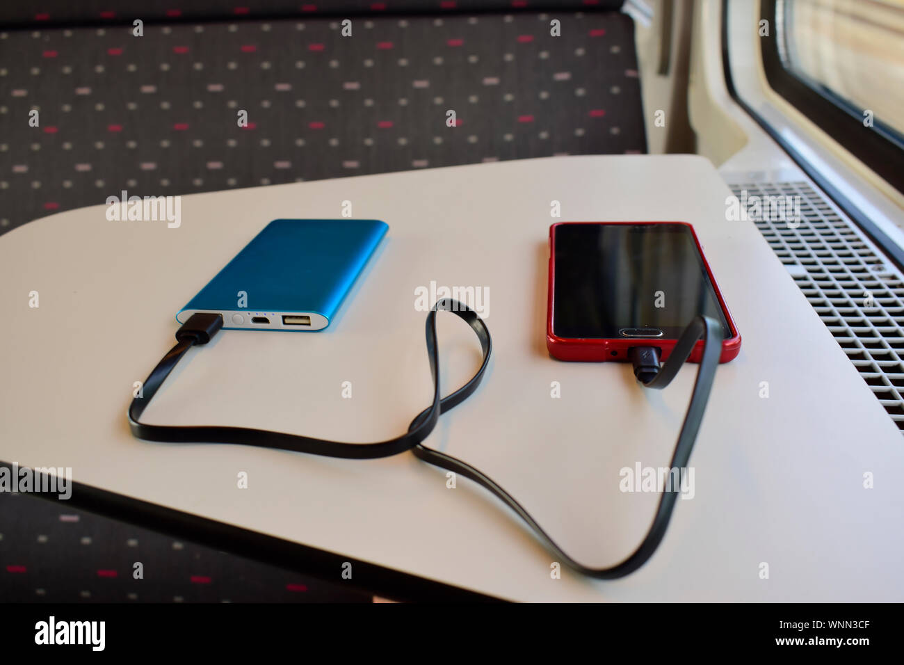 Schwarz und Rot Smartphone laden mit Blue Power Bank auf weißem Hintergrund in der Nähe des Sitzes im Zug gegen die Sonne durch das Fenster. Stockfoto