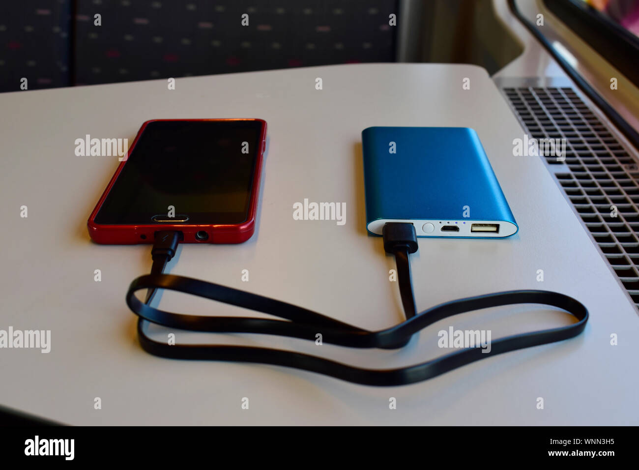 Schwarz und Rot Smartphone laden mit Blue Power Bank auf weißem Hintergrund in der Nähe des Sitzes im Zug gegen die Sonne durch das Fenster. Stockfoto