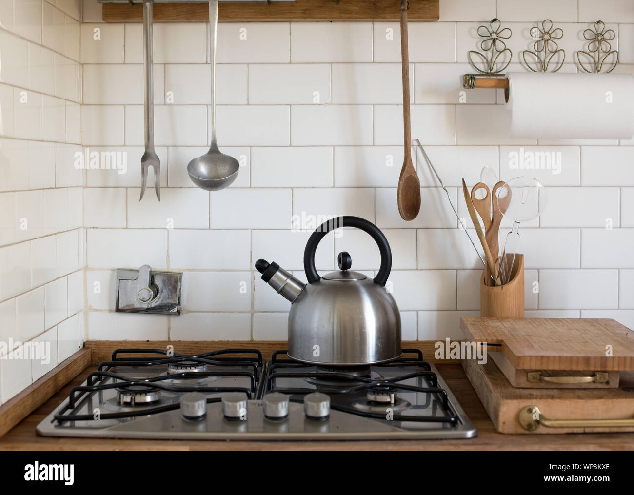 Edelstahl Wasserkocher stehen auf einem unbeleuchteten Gasbrenner auf dem Herd in der Küche mit Fliesen- white Splash Back und herabhängenden Utensilien Stockfoto