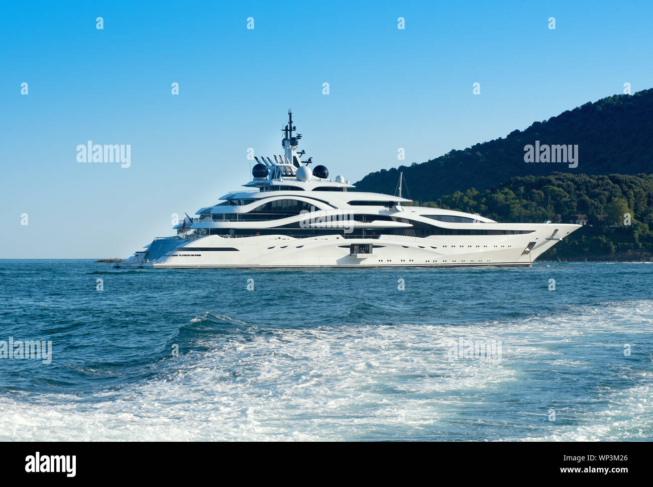Riesige Luxury Motor yacht offshore Cruising in einem ruhigen Meer an einem Küstenstreifen mit Wald über dem Schreibtisch eines vorbeifahrenden Schiff gesehen Stockfoto
