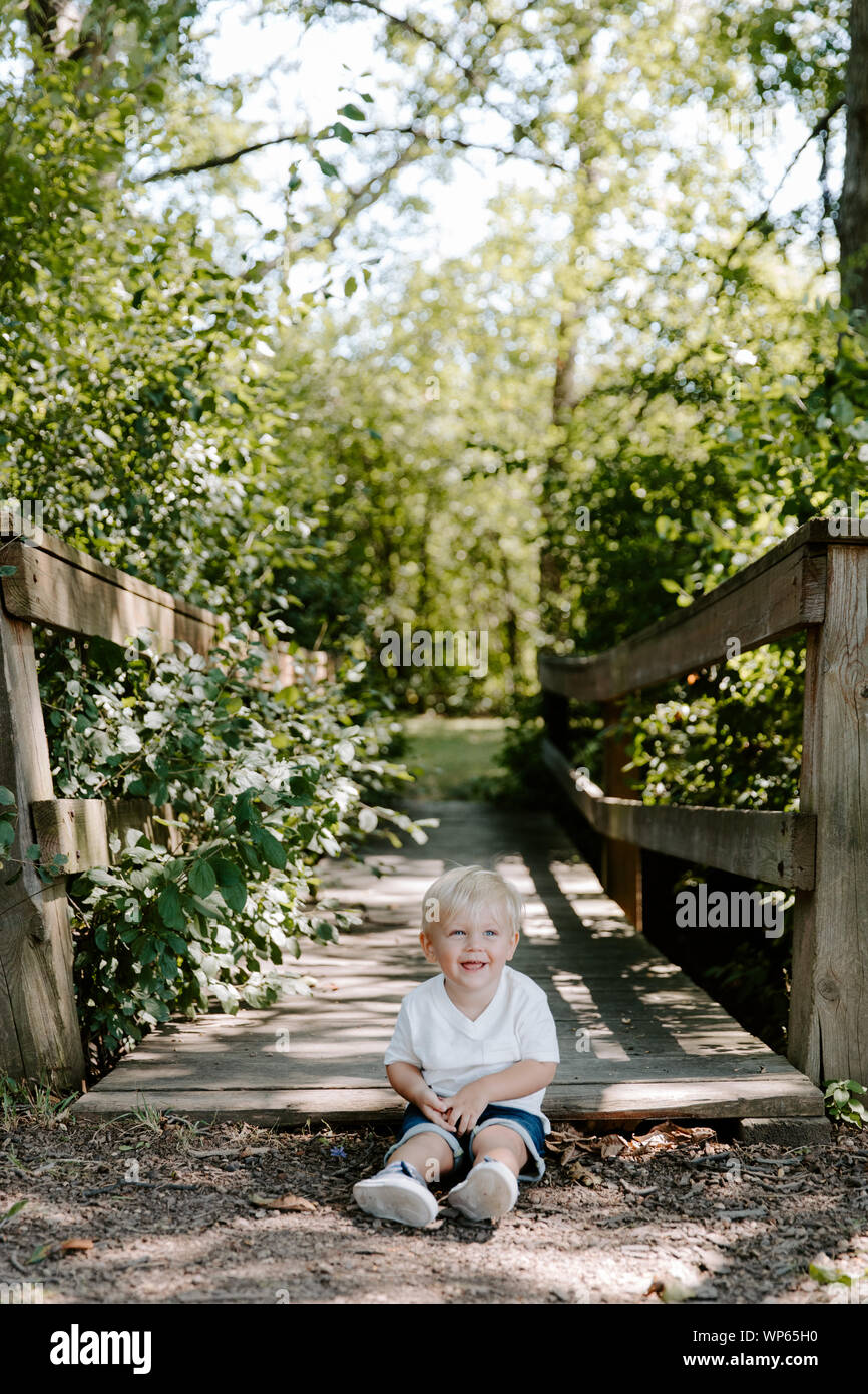 Süße kleine blonde Kind Junge Kind sitzen und Lachen vor der hölzernen Brücke über einen Bach im Park im Wald im Sommer Stockfoto