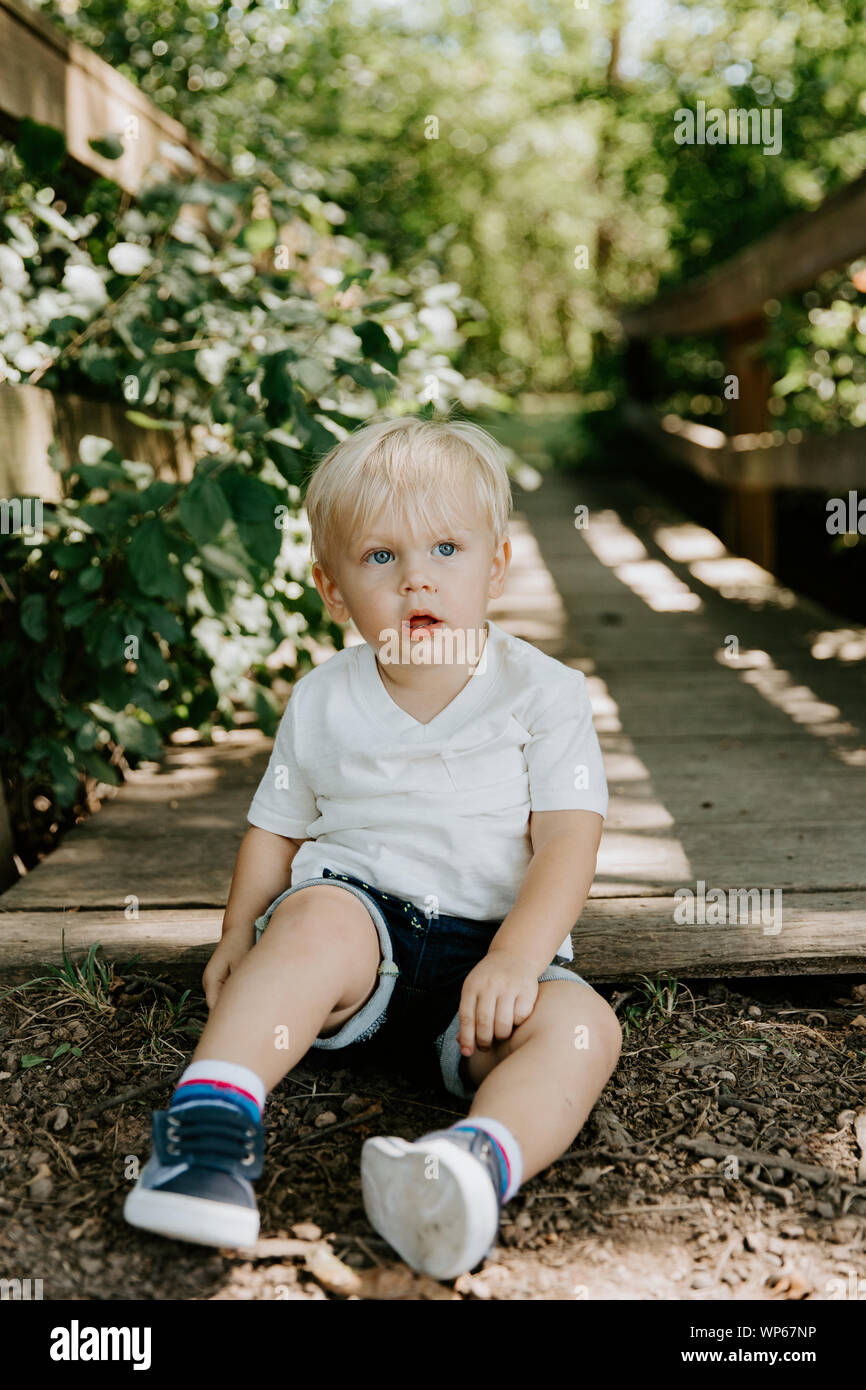 Süße kleine blonde Kind Junge Kind sitzen und Lachen vor der hölzernen Brücke über einen Bach im Park im Wald im Sommer Stockfoto