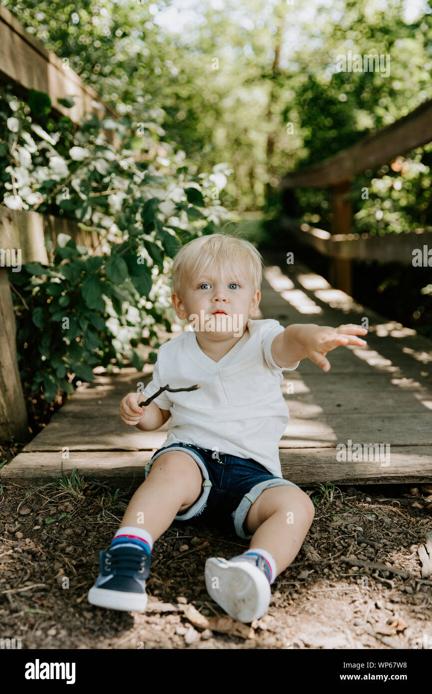 Süße kleine blonde Kind Junge Kind sitzen und Lachen vor der hölzernen Brücke über einen Bach im Park im Wald im Sommer Stockfoto