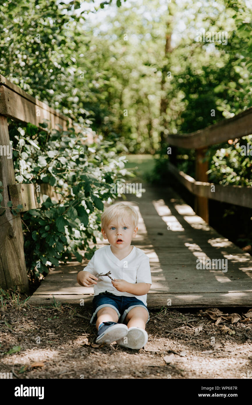 Süße kleine blonde Kind Junge Kind sitzen und Lachen vor der hölzernen Brücke über einen Bach im Park im Wald im Sommer Stockfoto