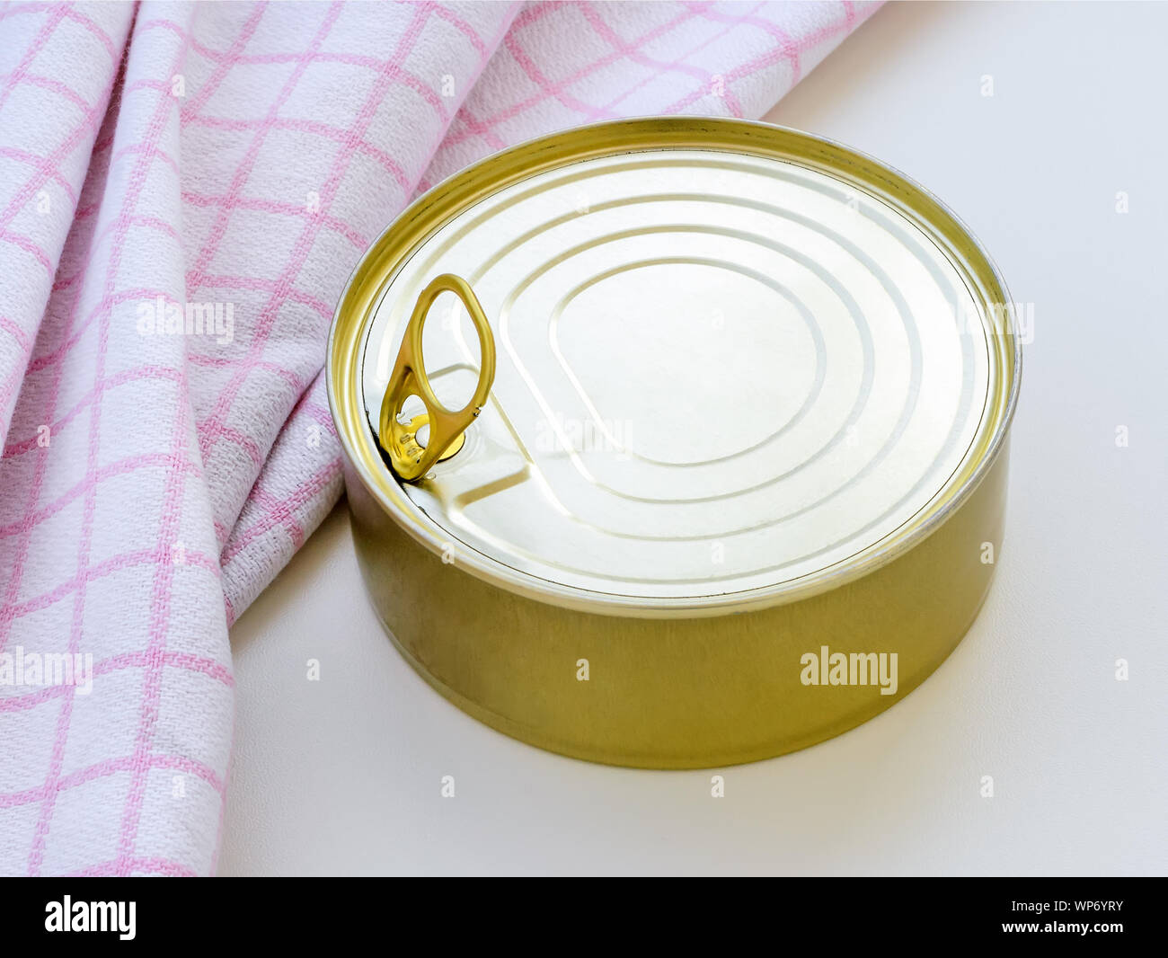 Gelber runder Blechdose in der Nähe von karierten Küchenhandtuch auf einem weißen Tisch. Öffnen einer Essen kann mit Lasche ziehen. Close-up. Stockfoto
