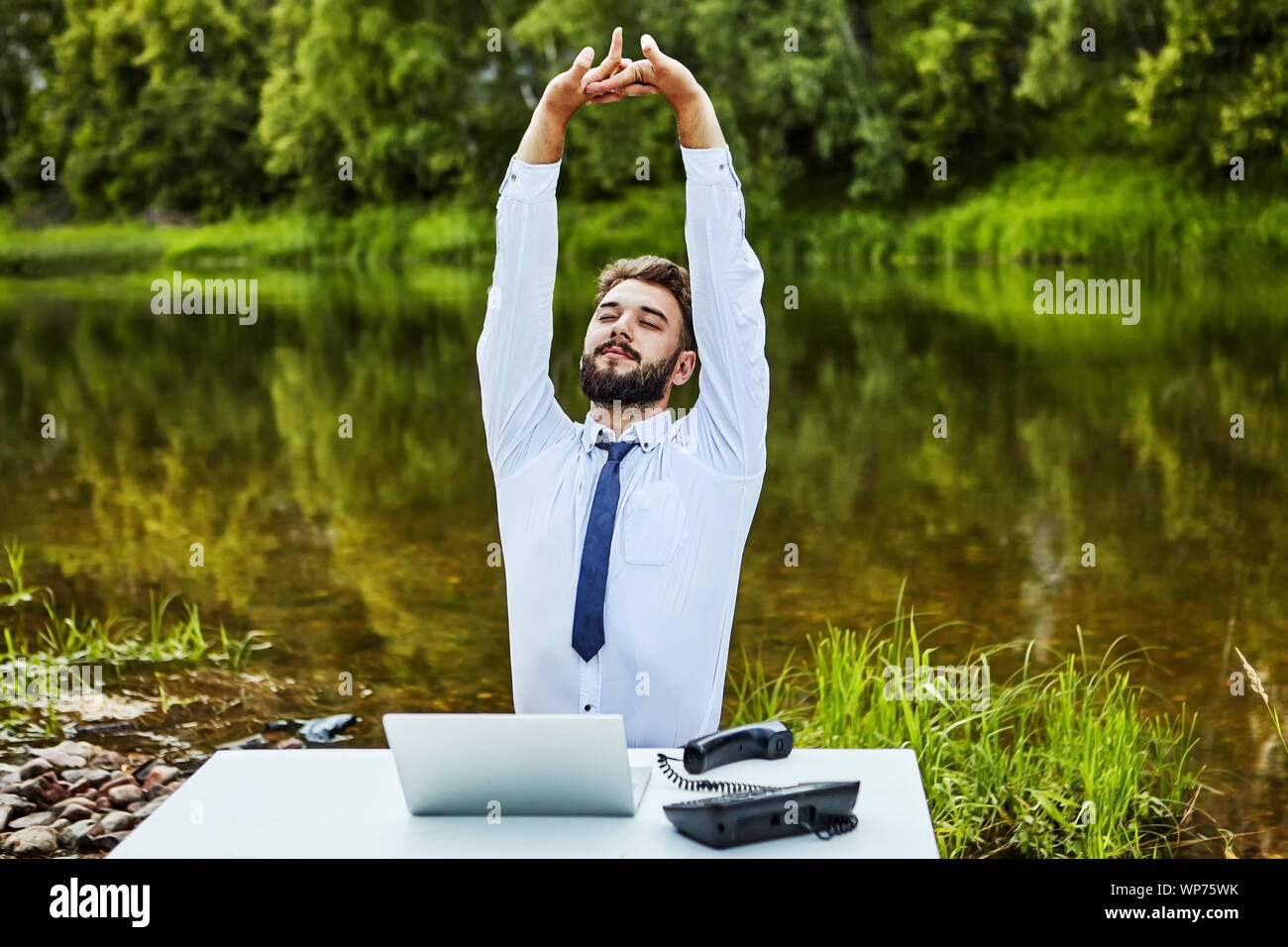 White Collar erstreckt sich beim Sitzen am Tisch. Ein Büroangestellter sitzt an einem Tisch inmitten einer natürlichen Landschaft mit einem wilden Fluss. Auf seinem Schreibtisch ist ein Erl Stockfoto