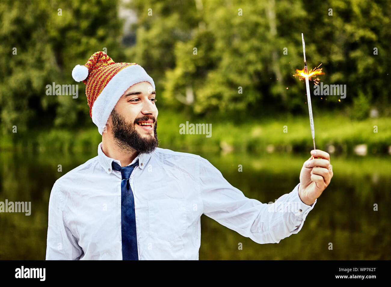 Ein Geschäftsmann in einem lustigen Weihnachtsmann Mütze schaut auf eine brennende Wunderkerze in der Hand. Sein Büro feiert das neue Jahr, oder Weihnachten. Eine Wunderkerze brennt in Th Stockfoto