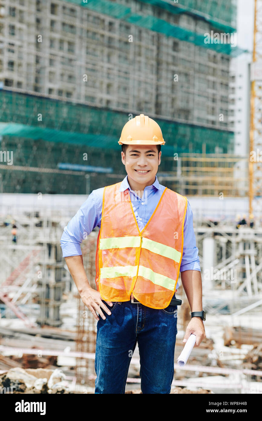 Portrait von positiven Vietnamesischen Kopf Ingenieur in helle Weste mit blueprint Blättern auf der Baustelle Stockfoto