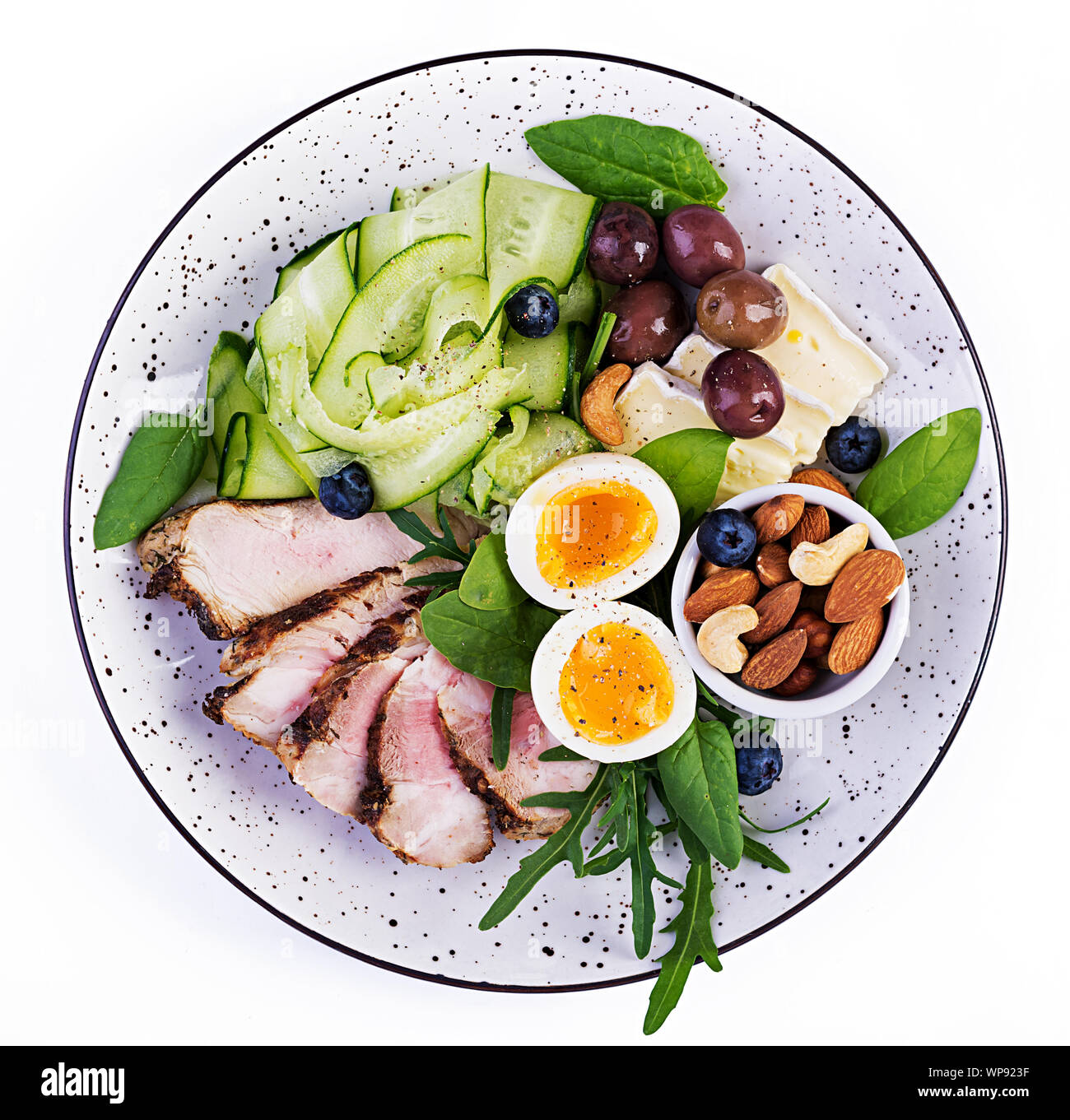 Ketogenic Diät. Keto Brunch. Gekochte Eier, Schweinesteak und Oliven, Gurken, Spinat, Brie Käse, Nüsse und Heidelbeeren. Ansicht von oben Stockfoto