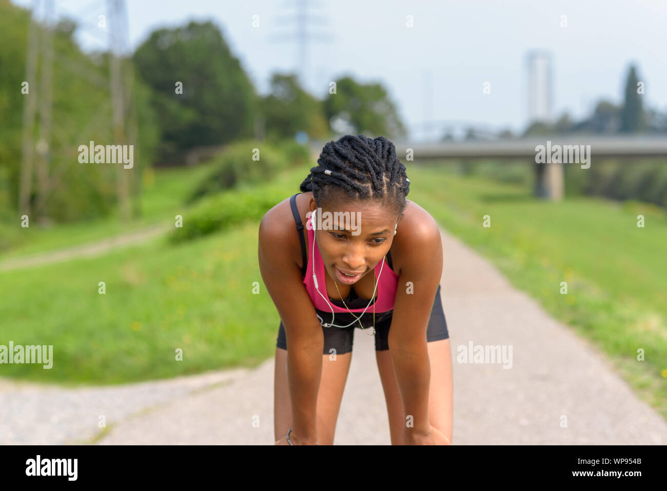 Junge athletische afrikanisches Mädchen eine Pause vom Joggen nach vorn auf die Knie gelehnt verspannt ruht während der Wiedergabe von Musik über Ohrstöpsel Stockfoto